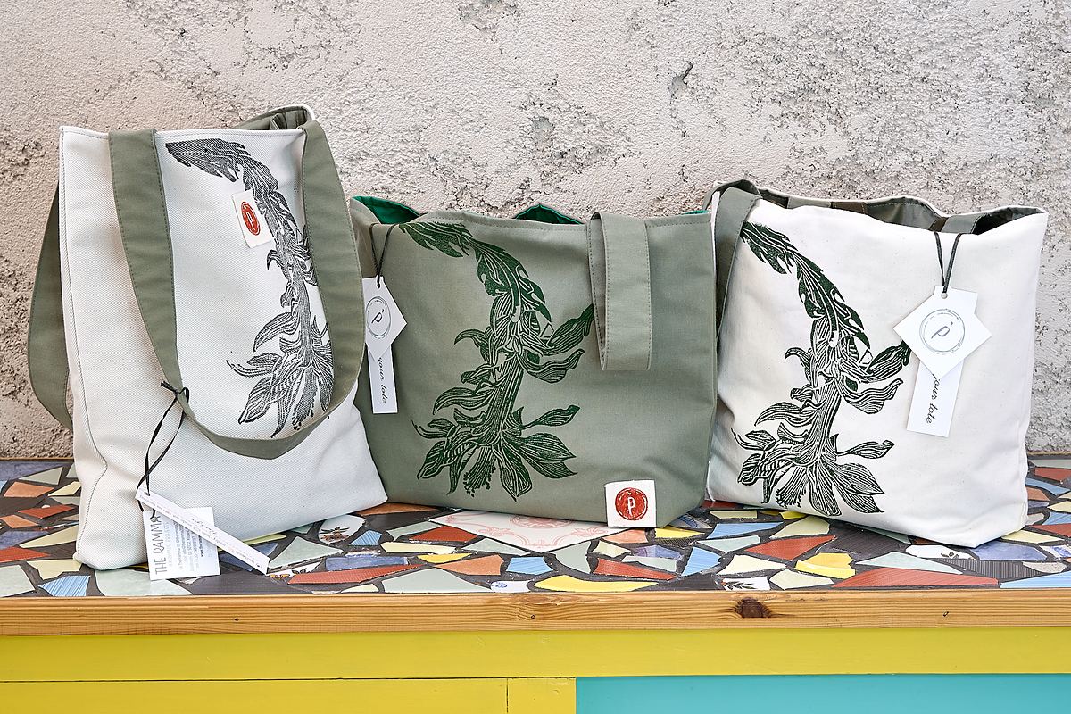 Three canvas totes in sage and natural cotton lined up on a mosaic bench, each printed with a different hand-carved botanical block.
